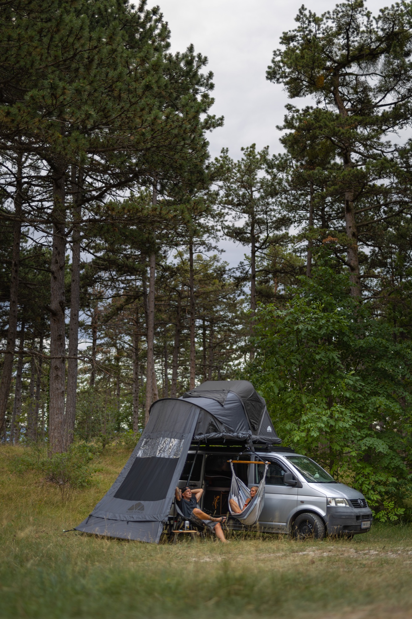 Paar entspannt in Hängematte und Campingstuhl unter dem Vorzelt am VW-Bus im Kiefernwald