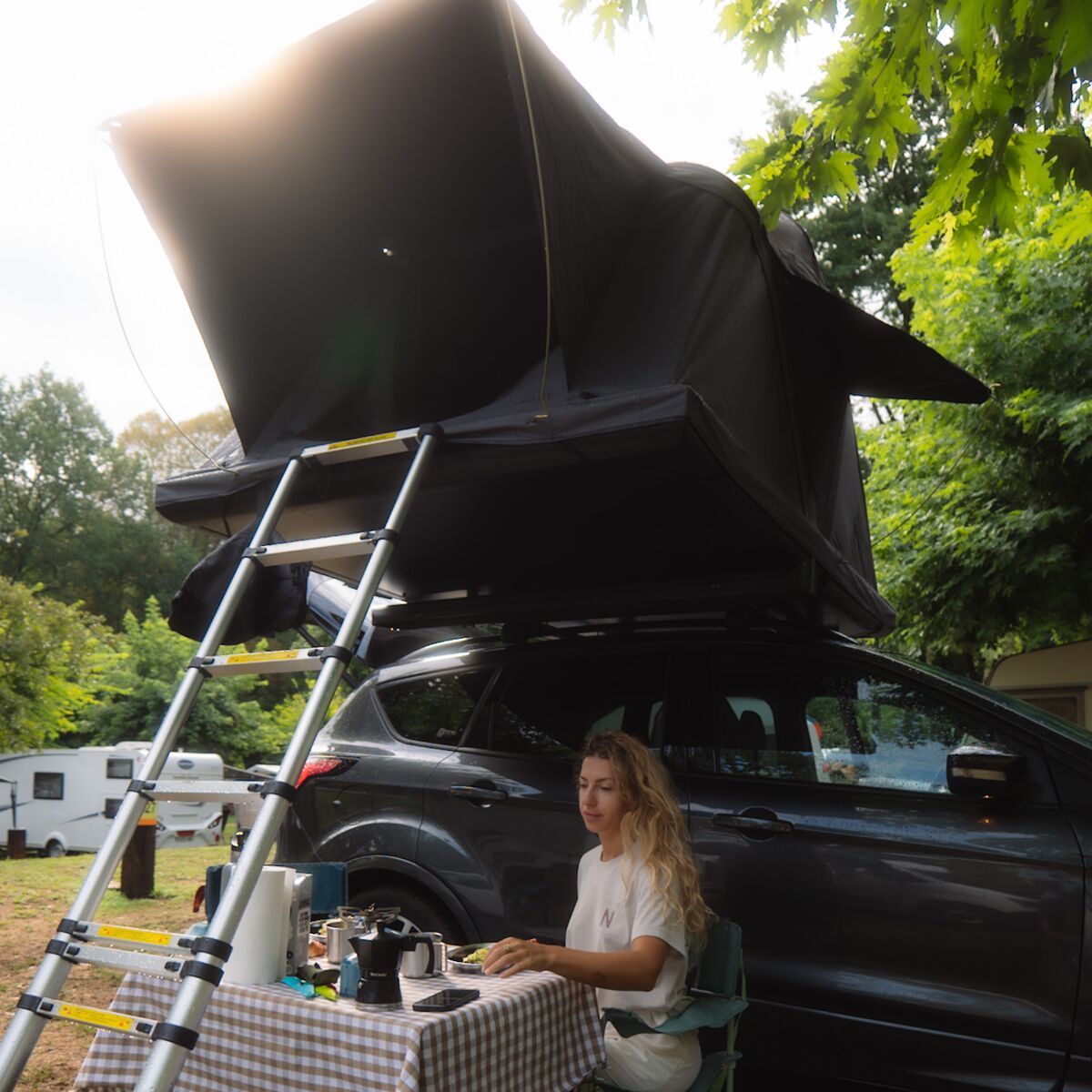 Frau am Campingtisch neben einem Auto mit geöffnetem Roof Space 2 Dachzelt im Wald